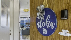 A wooden door with a 'aloha' sign, a small black device, and a view into a living room with a sofa and a lamp. - Stunning Oceanfront Retreat, Kahana Reef 218  (Lahaina)