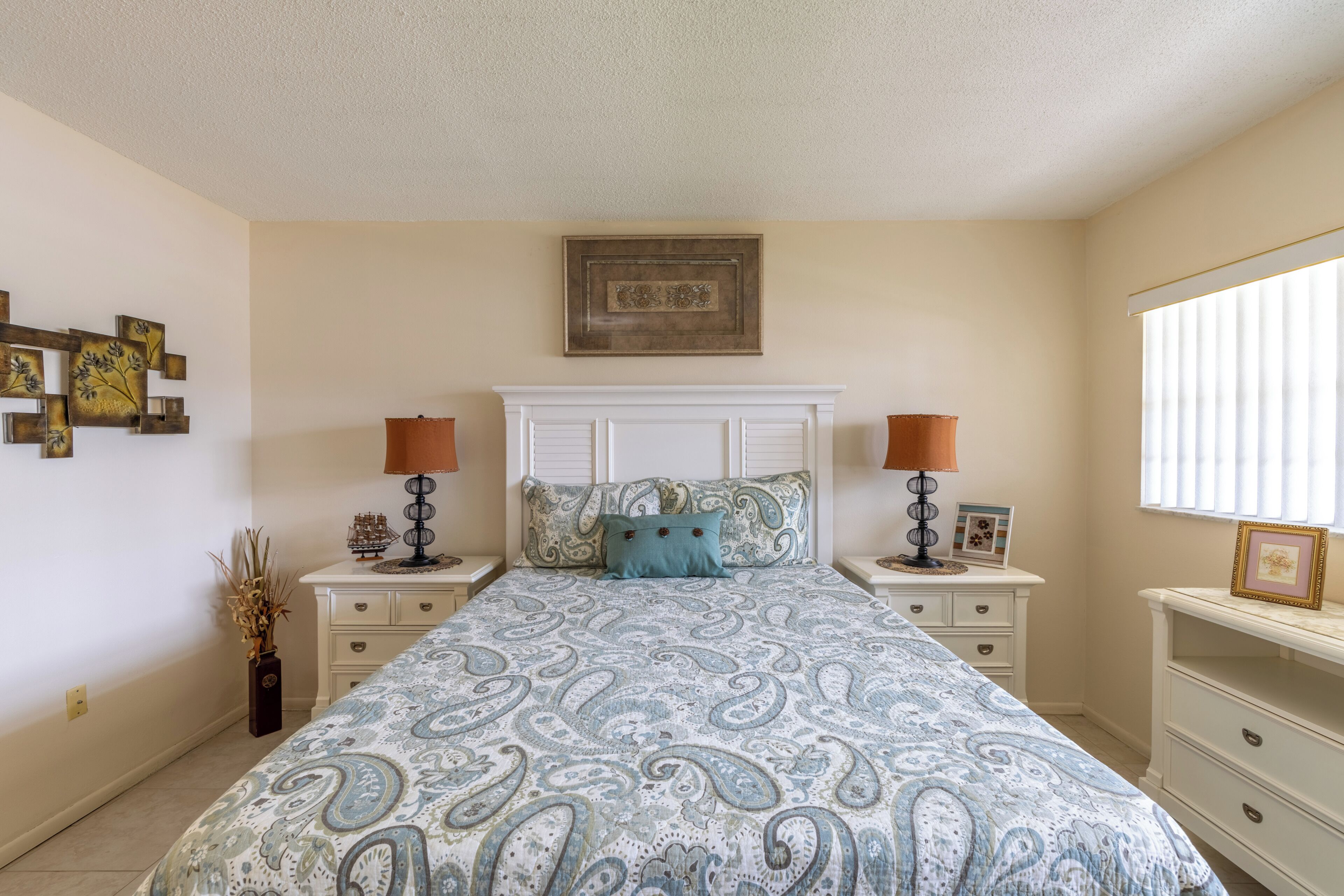 A bedroom with a patterned bedspread, two bedside tables with lamps, a nightstand, a dresser, and a wall-mounted picture frame.