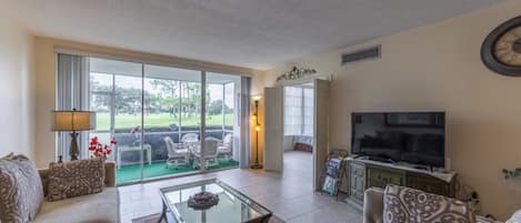 A spacious living room with a large television, comfortable seating, and a view of a green landscape through the glass doors.