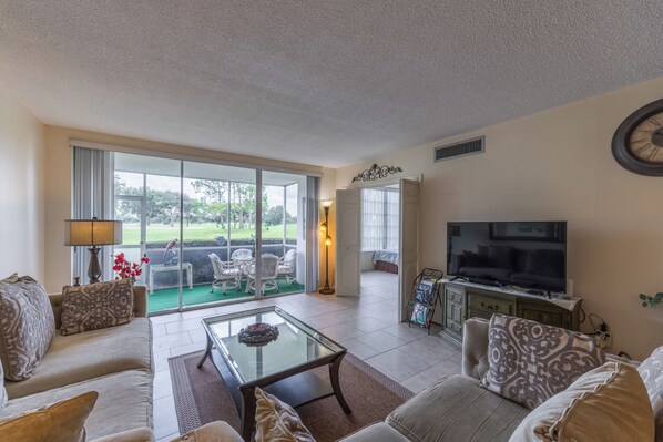A spacious living room with a large television, comfortable seating, and a view of a green landscape through the glass doors.