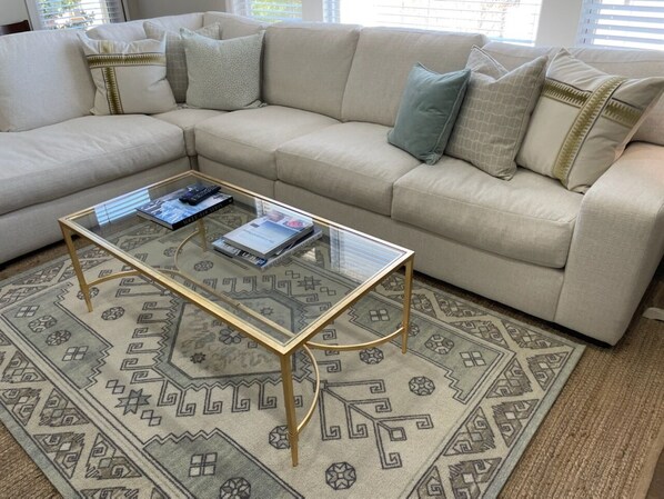 Ein Wohnzimmer mit einem beigefarbenen Ecksofa, einem Glastisch mit Büchern und einem gemusterten Teppich.