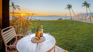 A table with a pineapple, a bottle, and a glass on a glass table, overlooking a grassy area with palm trees and a view of the ocean at sunset. - 104 - Lovely Ground Floor Direct Oceanfront + Larger Lanai (Kihei)