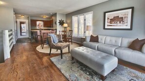 A spacious living room with a grey sofa, a round coffee table, and a patterned rug. A kitchen is visible in the background with a dining area and bar stools. - GREAT OLD TOWN LOCATION- SKI TO TOWN LIFT & STEPS TO MAIN ST!  (Park City)