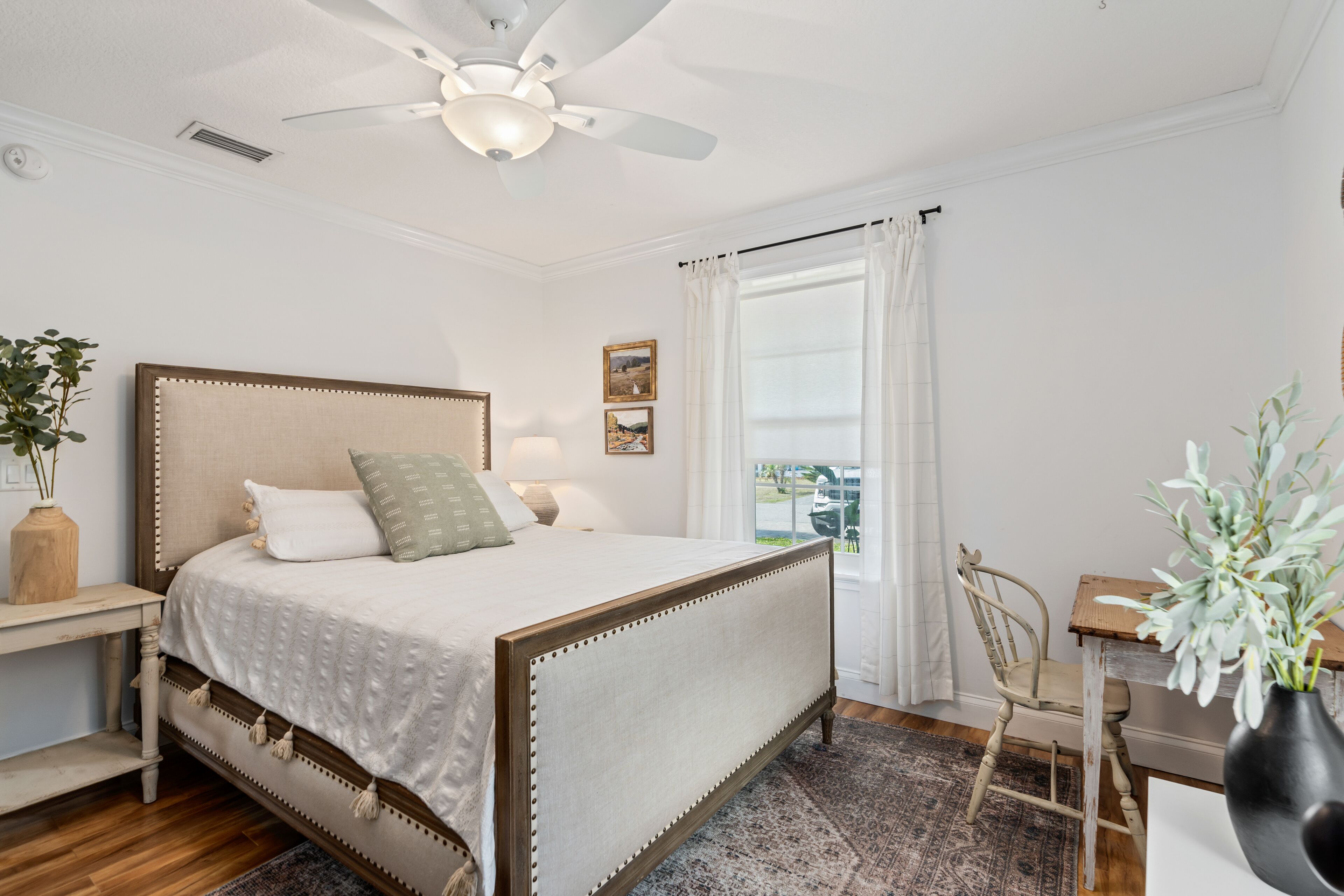 A bedroom with a bed, a ceiling fan, a window with curtains, a chair, and a small table with a vase.