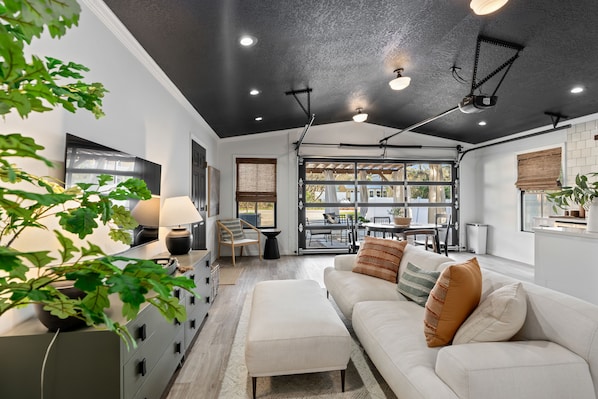 Uma sala de estar moderna com um sofá branco, uma mesa de centro e uma área de jantar com mesa e cadeiras. Há uma janela grande que oferece luz natural e vista para o exterior.