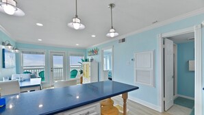 A kitchen with a blue countertop, white cabinets, and wooden stools. There are pendant lights, a window, and a door leading to another room. - Stunning Beachfront Condo - Recently Remodeled! (Galveston)