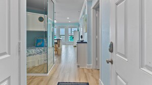 A hallway with a glass door leading to a bedroom with a bunk bed, a kitchen area with a counter and stools, and a view of the outdoors through a window. - Stunning Beachfront Condo - Recently Remodeled! (Galveston)