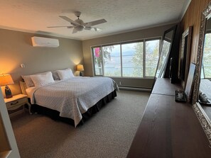 Une chambre à coucher comprenant un lit, une table de chevet, une lampe, un ventilateur de plafond, une grande fenêtre donnant sur l’extérieur et une commode avec un miroir.