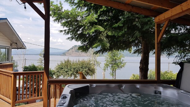 Une terrasse en bois avec un jacuzzi, donnant sur un lac, avec des arbres et des montagnes en arrière-plan.