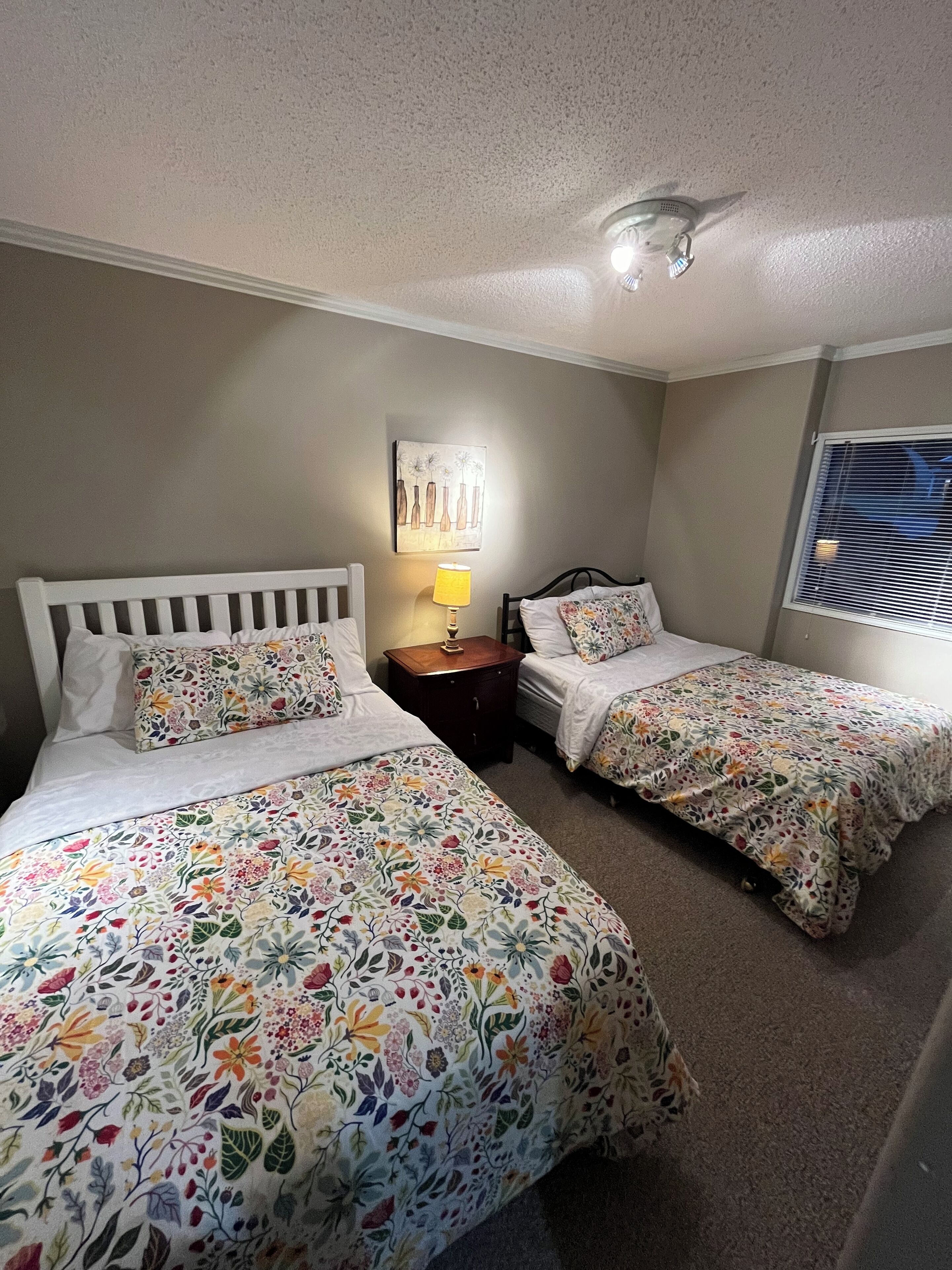 A room with two single beds, a nightstand with a lamp, a wall-mounted artwork, and a window with blinds.