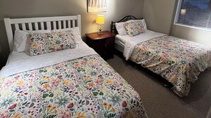 Une chambre avec deux lits simples, une table de chevet avec une lampe, une œuvre d’art accrochée au mur et une fenêtre avec des stores.
