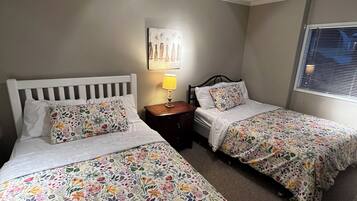 A room with two single beds, a nightstand with a lamp, a wall-mounted artwork, and a window with blinds.