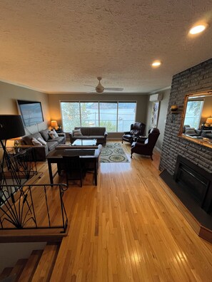 A spacious living room with a fireplace, a television mounted on the wall, and a large window offering a view of the outdoors.