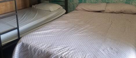 Una cama litera con una base inferior y una cama individual, ambas con ropa de cama sencilla. Se ve un armario de madera a la izquierda.