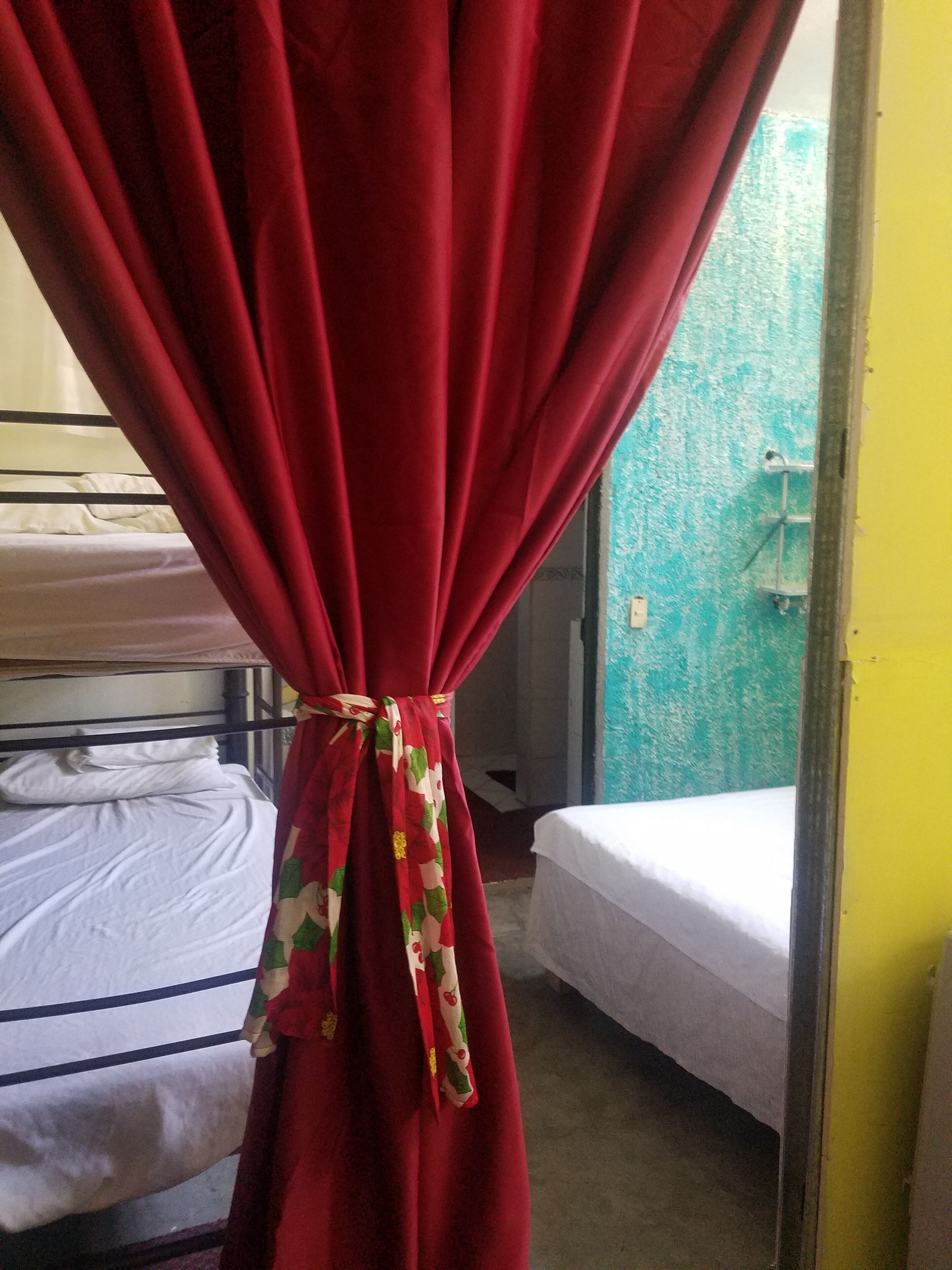 Quarto com cortina vermelha amarrada na parte inferior, uma cama de solteiro com roupa de cama branca e uma cama individual com roupa de cama branca.
