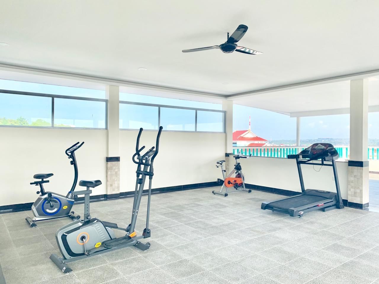 Un gimnasio con varios equipos de ejercicio, ventanas amplias y un ventilador de techo.