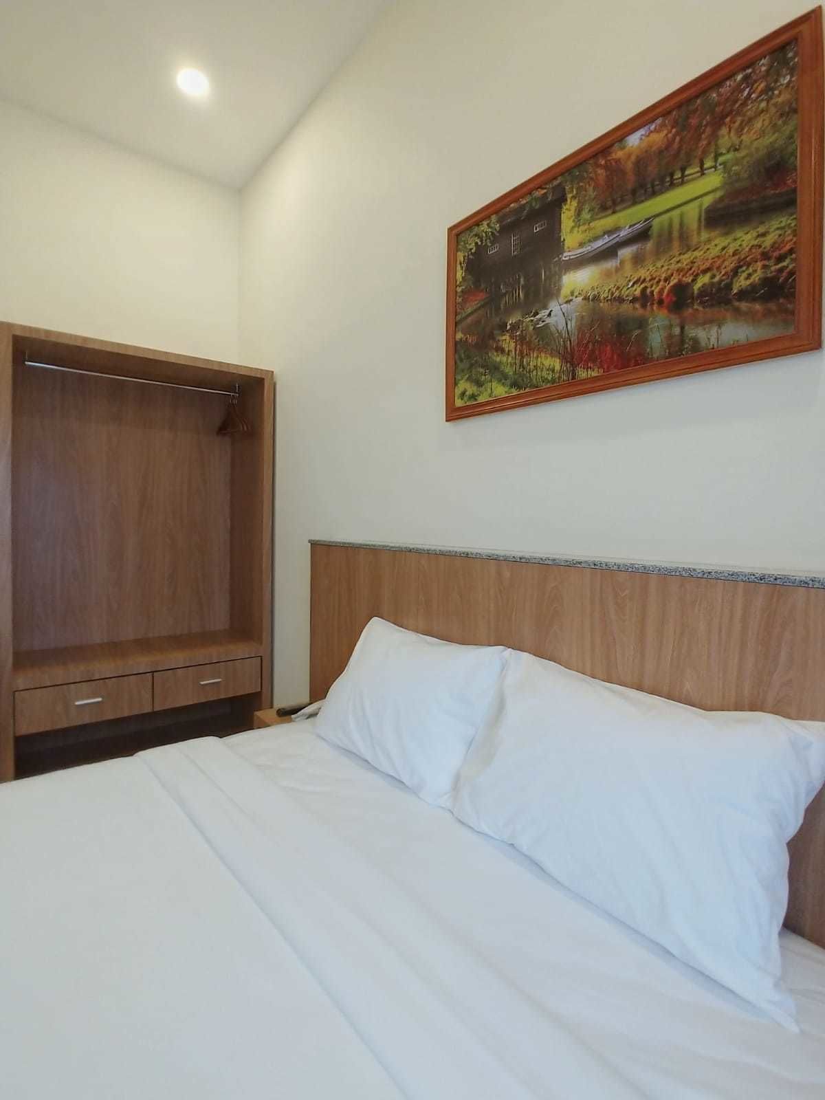 A bedroom with a wooden headboard, a bed with white bedding, a wooden wardrobe, and a painting of a landscape on the wall.
