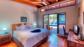 A bedroom with a bed, a desk with a computer, a TV, and a view of an outdoor pool area. - Beachfront Private Villa (Korolevu)