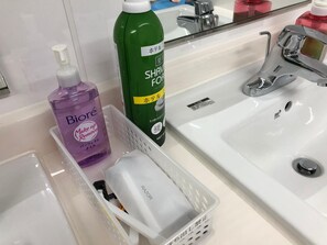 A bathroom with a white sink, a mirror, and toiletries including a bottle of Biore makeup remover, a bottle of Shave Soap, and a razor in a basket. - Hotel Mirai (Yokohama)