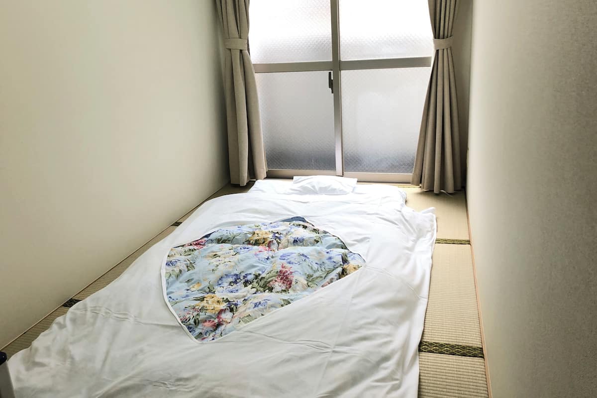 Una habitación de hotel estrecha con una cama, una ventana con cortinas y vistas al exterior.