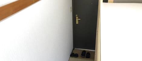 A narrow hallway with a wooden bench on the right, a door with a doorknob, and a small shoe rack with shoes.