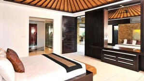 A modern bedroom with a large bed, a wooden ceiling, a dark wall panel, and a bathroom visible through an open door. - Modern Family Villa (Kerobokan)