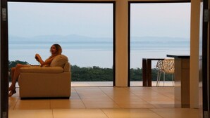 A person sitting on a sofa by a large window with a view of a body of water and mountains. - Fiji Luxury Pool Villa (Lautoka)