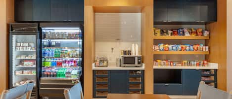 Eine kleine Küchenzeile mit Kühlschrank, Mikrowelle und Regalen voller Snacks und Getränke.