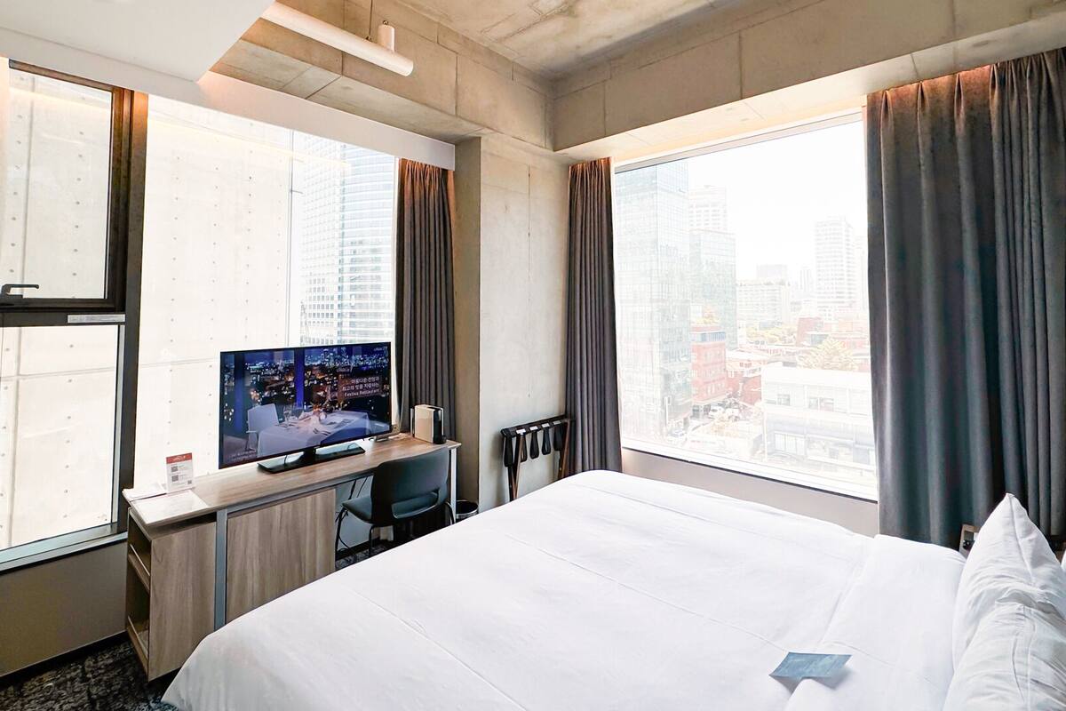 Ett hotellrum med en stor säng, ett skrivbord med en dator, en tv och utsikt över staden genom stora fönster.