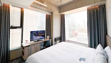 Habitación de hotel con una cama grande, un escritorio con computadora, un televisor y vistas a la ciudad a través de ventanas amplias.