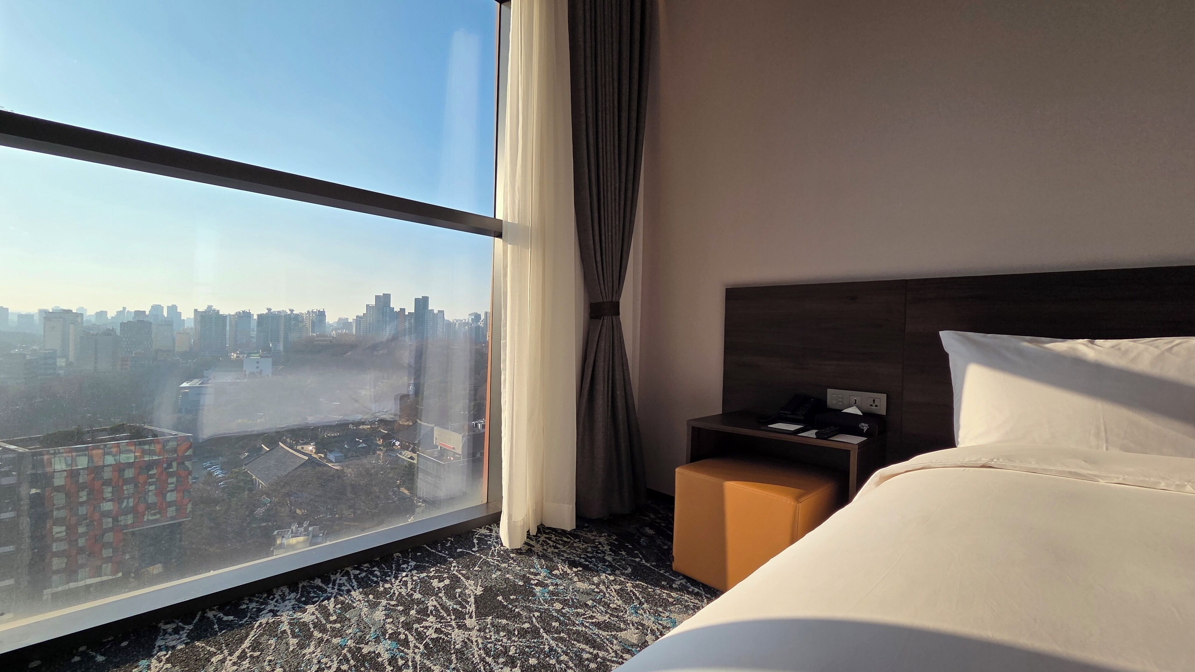 Ein Hotelzimmer mit großem Fenster, einem Bett, einem Nachttisch und Blick auf eine Stadtlandschaft.