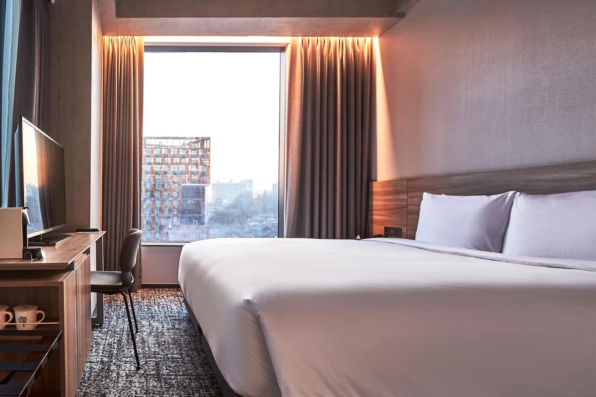 Ett hotellrum med en stor säng, ett skrivbord med en stol, en TV och utsikt över stadsbilden genom fönstret.