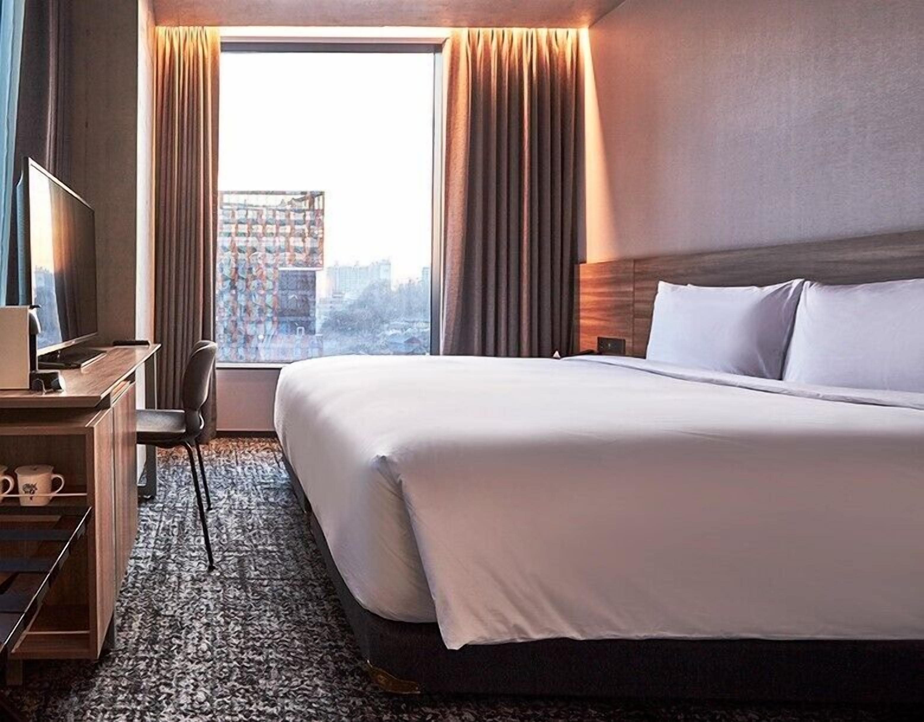 Una habitación de hotel con una cama grande, un escritorio con computadora, una silla y vistas a la ciudad desde la ventana.