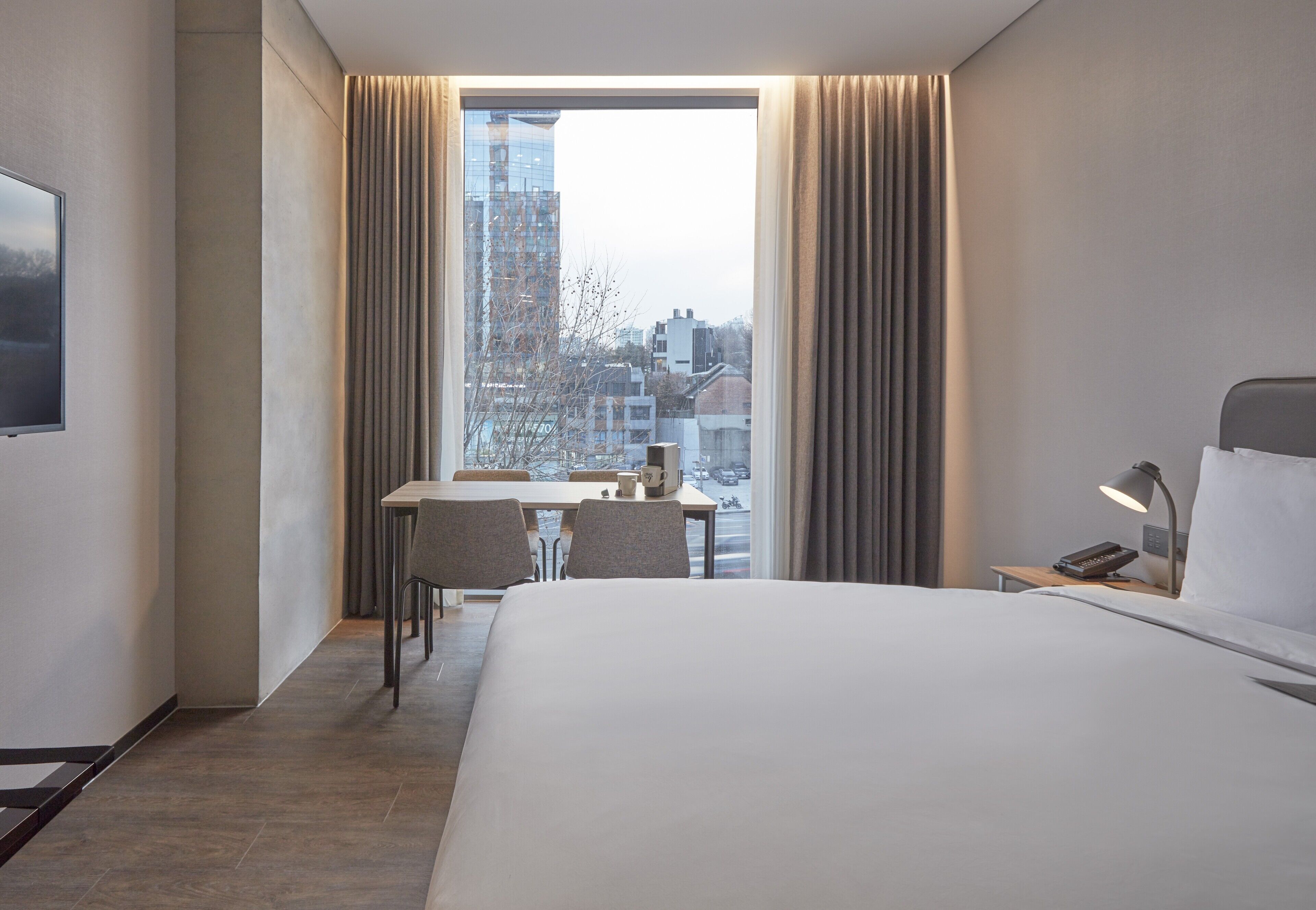A modern hotel room with a large bed, a desk with chairs, and a view of the city through a large window.