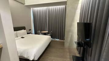 A hotel room with a large bed, a TV mounted on the wall, a small table, and a window with curtains.