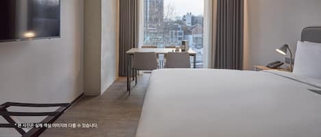 A modern hotel room with a large bed, a desk with a chair, a TV mounted on the wall, and a window with curtains.