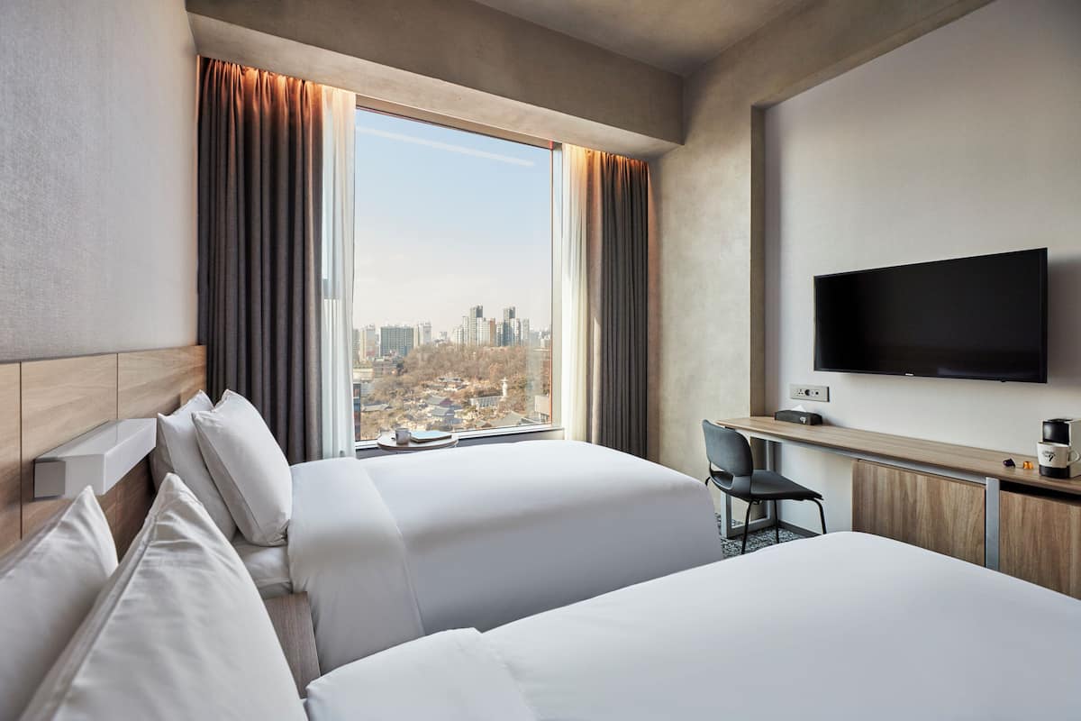 Ett hotellrum med två sängar, ett skrivbord, en stol, en TV och utsikt över staden.