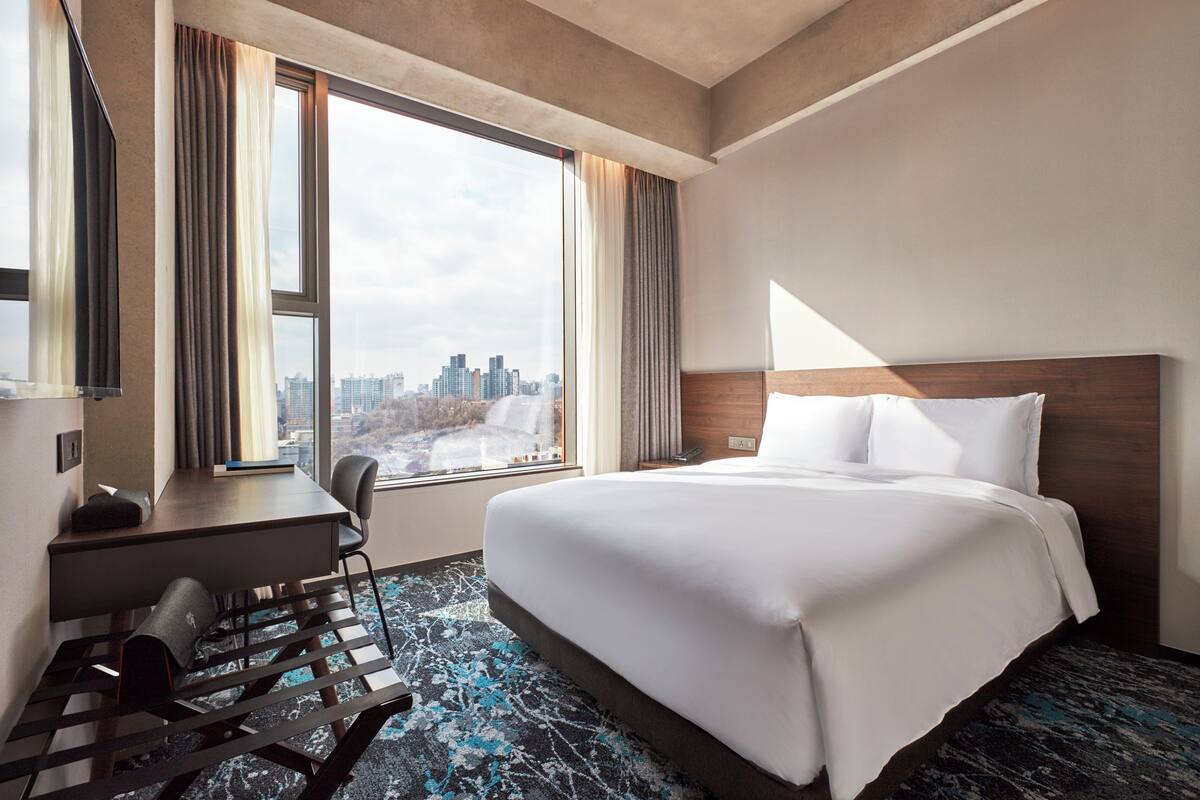 Ett hotellrum med en stor säng, ett skrivbord med en stol, utsikt över stadsbilden genom stora fönster och en modern mattdesign.