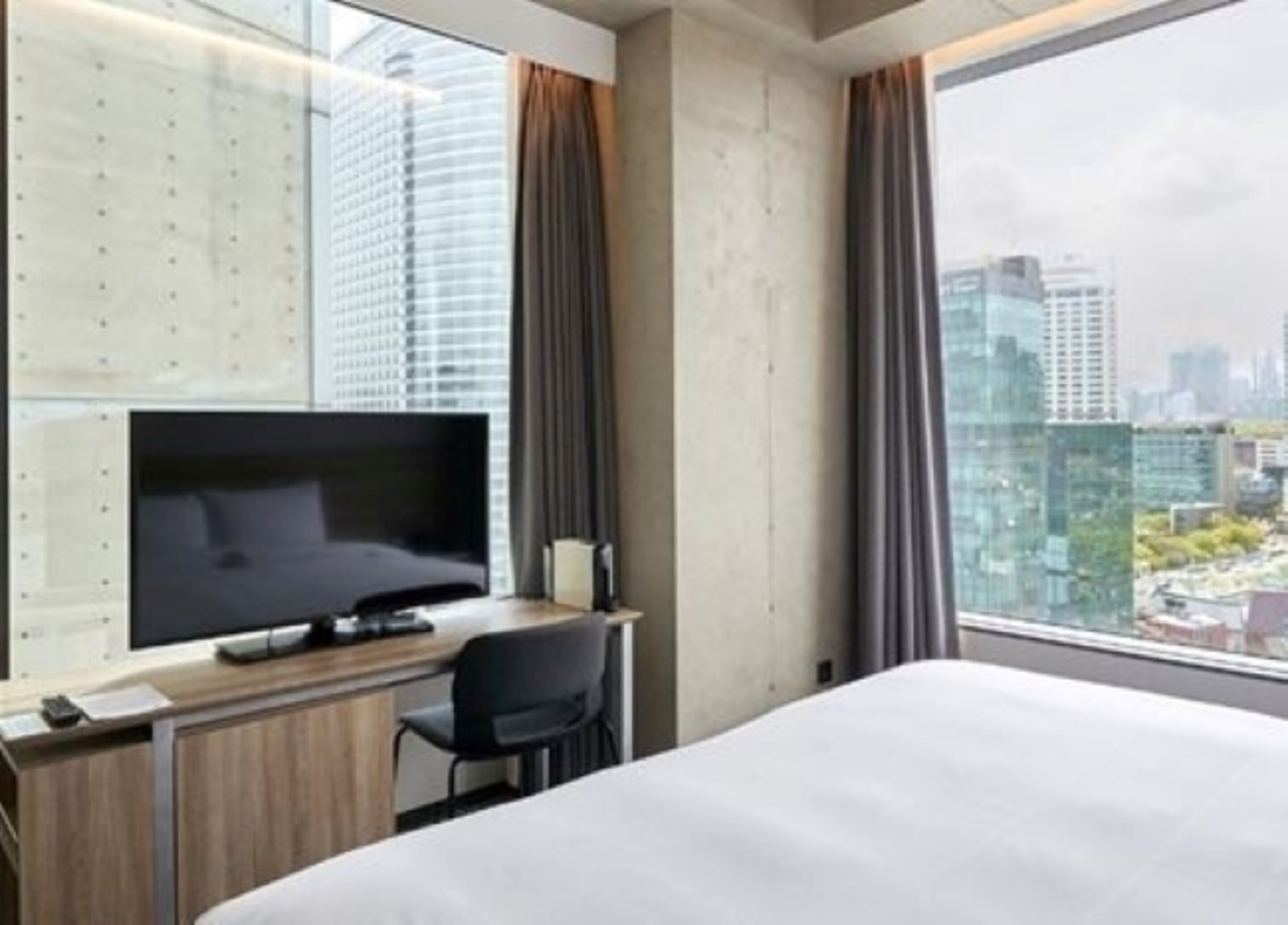 Una habitación de hotel moderna con una cama grande, un televisor de pantalla plana, un escritorio con una silla y vistas a la ciudad a través de amplias ventanas.