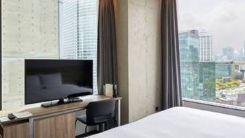 Una habitación de hotel moderna con una cama grande, un televisor de pantalla plana, un escritorio con una silla y vistas a la ciudad a través de amplias ventanas.