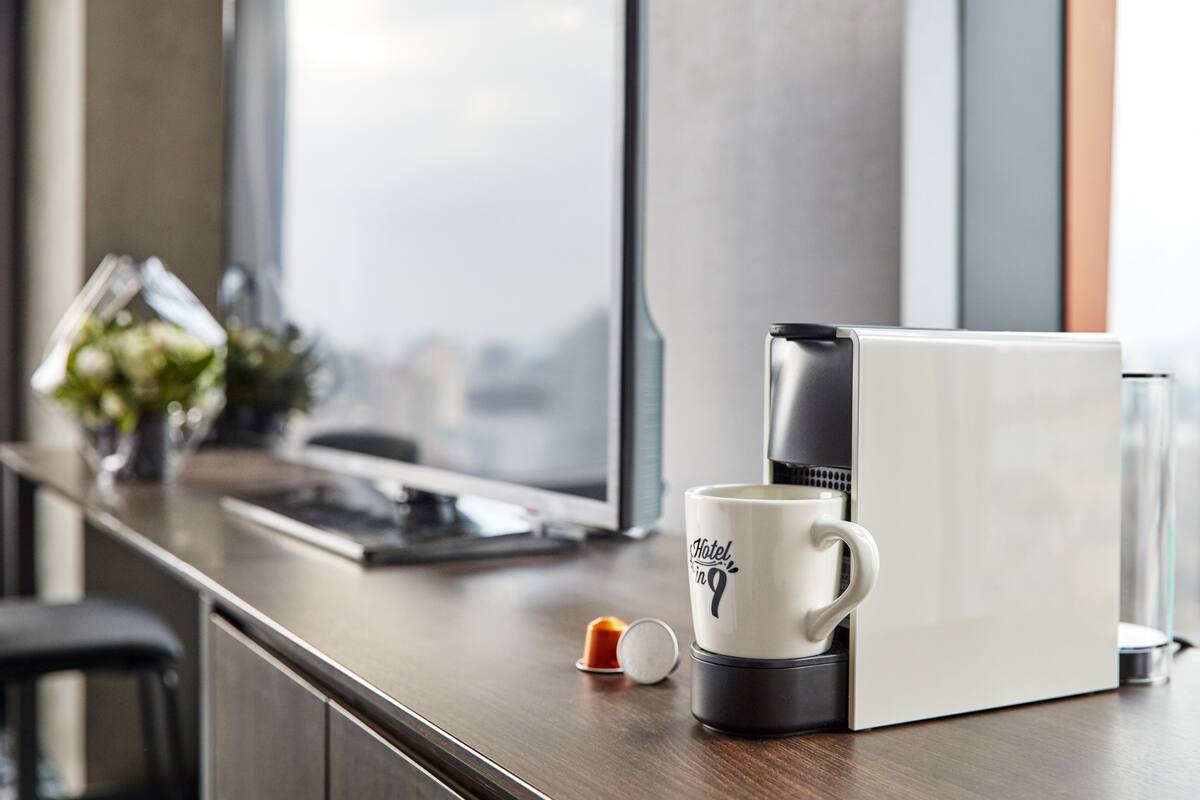 En kaffemaskin med en mugg på en träbänk, en liten växt och ett fönster med utsikt över staden.
