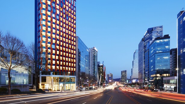 Un hotel moderno con una facciata importante nei colori rosso e blu, circondato da edifici alti e una strada trafficata con scie luminose che indicano il traffico.