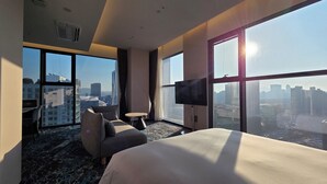 Una camera d'albergo moderna con un letto grande, una TV, un tavolino e un divano. La stanza ha ampie finestre con vista sulla città.