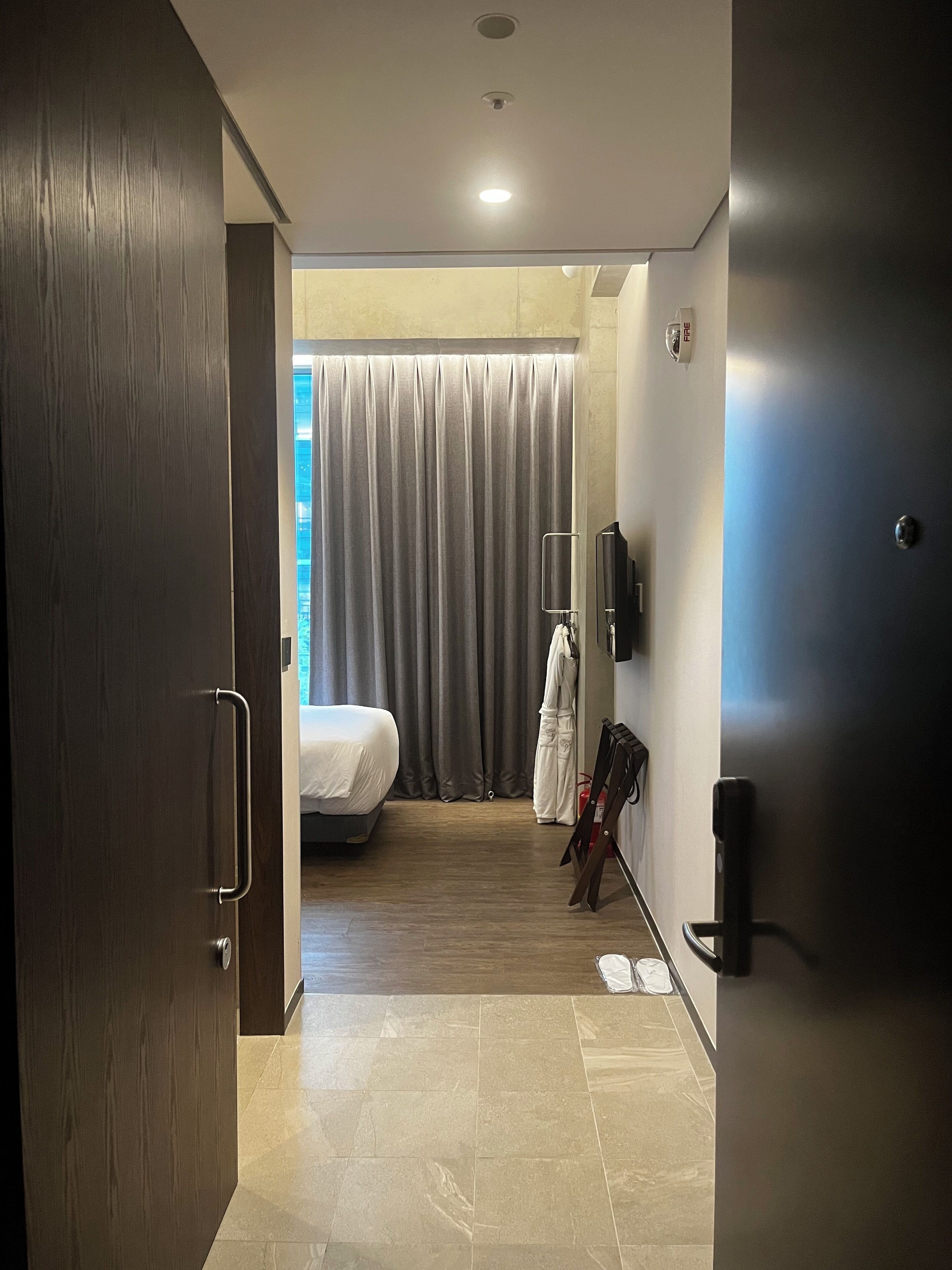 A hotel room with a bed, a wardrobe, a mirror, a fire alarm, and a curtain.