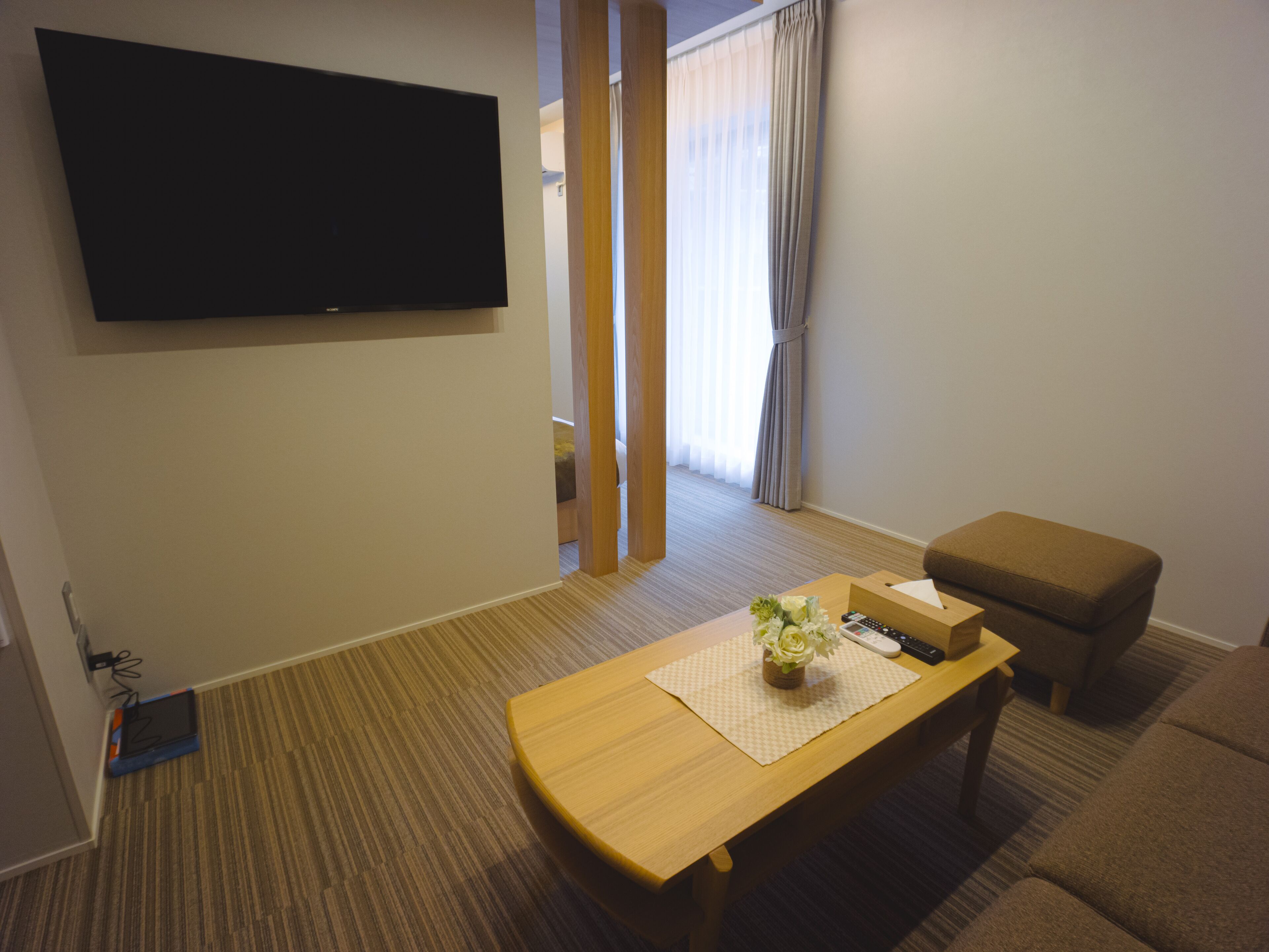 Habitación de hotel con televisión de pantalla plana en la pared, una mesa de centro de madera con un jarrón de flores, un sofá marrón y suelo de madera.