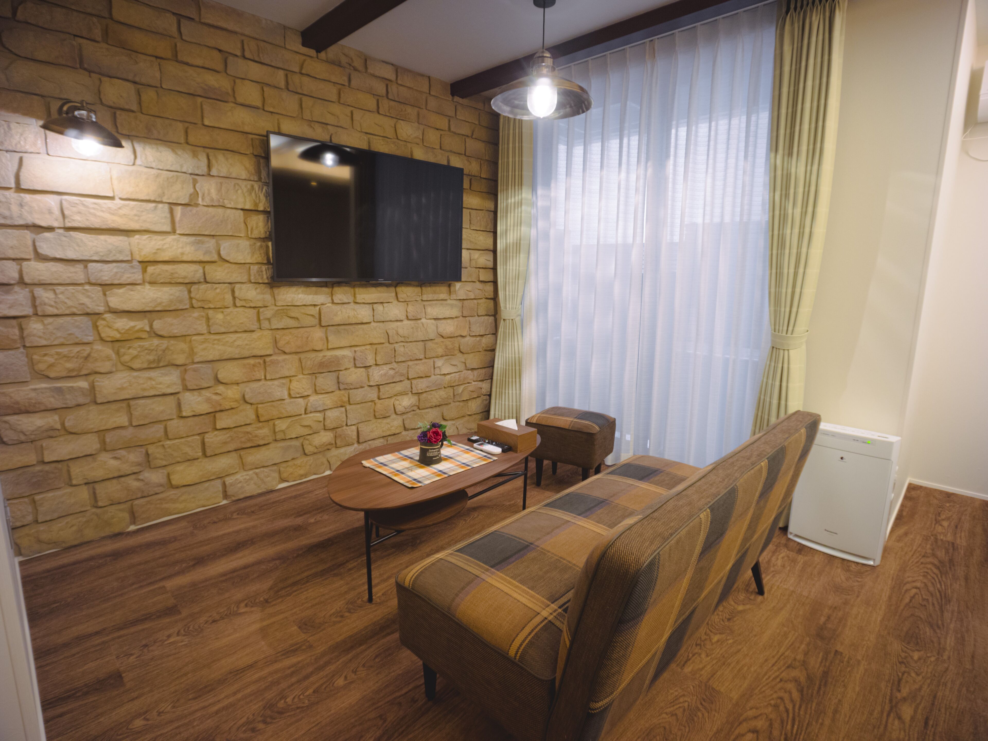 A cozy living room with a stone wall, a flat-screen TV, a wooden dining table, a plaid sofa, and a small table with a vase.