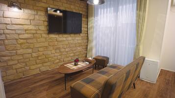 A cozy living room with a stone wall, a flat-screen TV, a wooden dining table, a plaid sofa, and a small table with a vase.