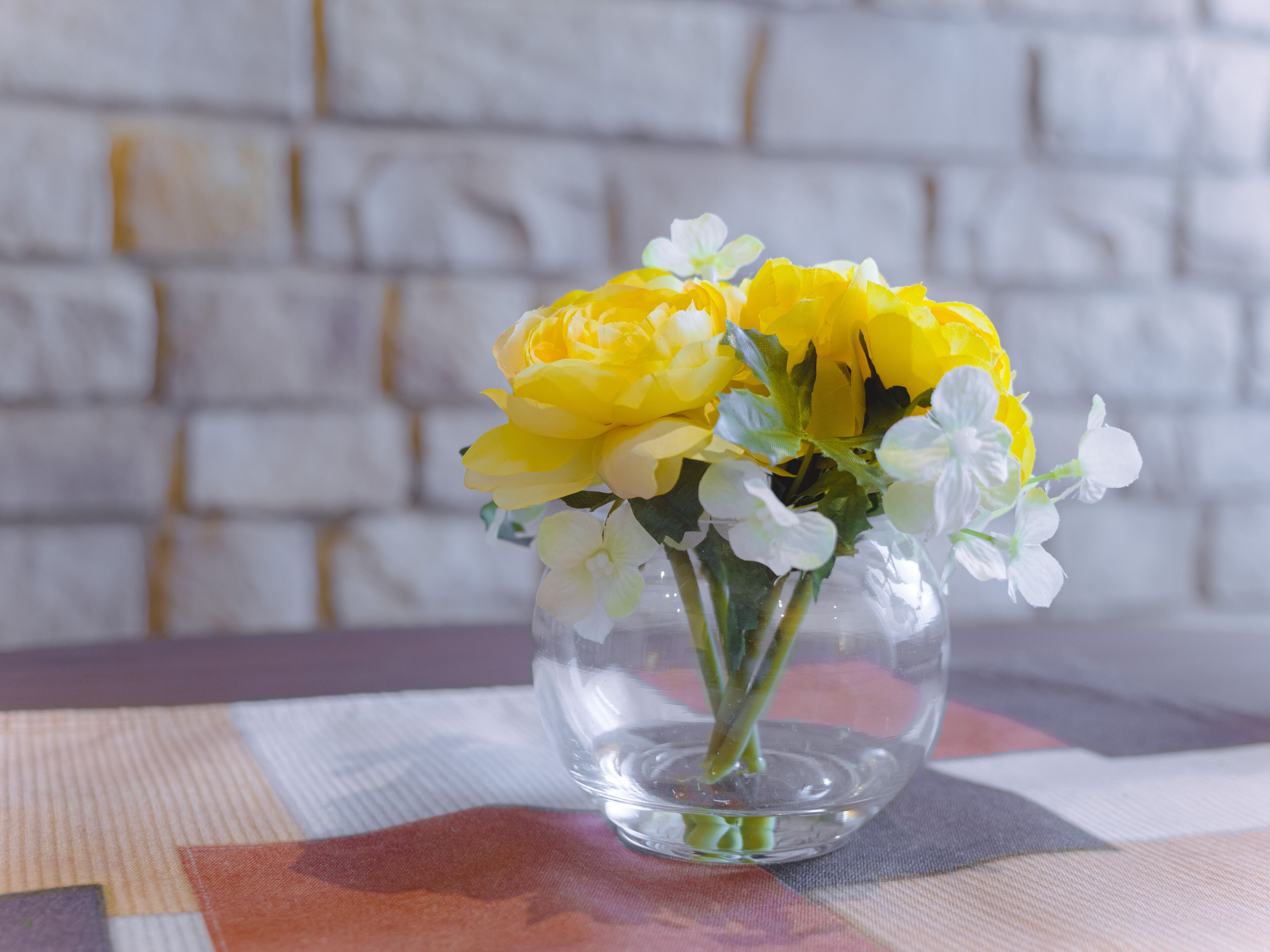 Un florero de vidrio transparente con flores amarillas y blancas sobre una mesa con mantel a cuadros.