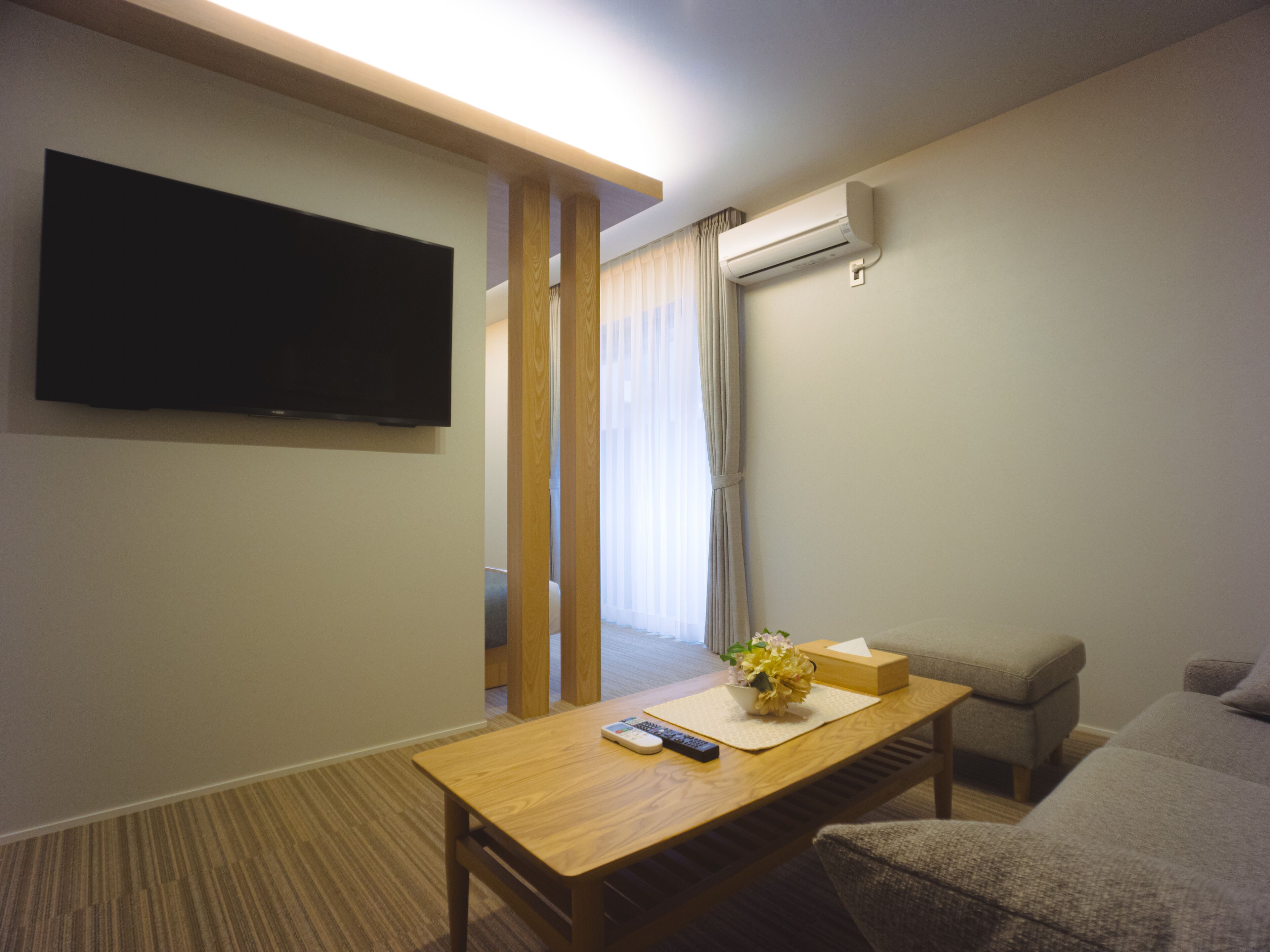 A hotel room with a flat-screen TV mounted on the wall, a wooden coffee table with a remote control and a vase of flowers, a grey sofa, and a window with curtains.