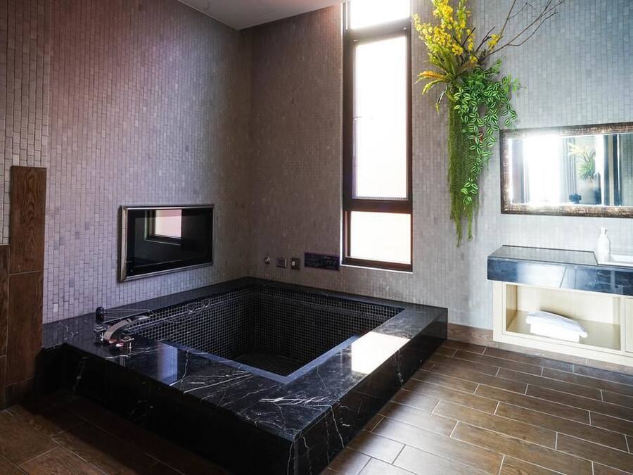 A modern bathroom with a large black marble bathtub, a built-in TV, and a large mirror above the sink.